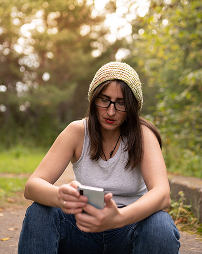 Lady wearing bobble hat, white vest top and jeans, sitting on a step in a park and looking at the her phone